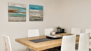 A dining area with a wooden table, white chairs, and two abstract paintings on the wall. - Beaches Apartment 13 – Bennett’s Beach Retreat (Hawks Nest)