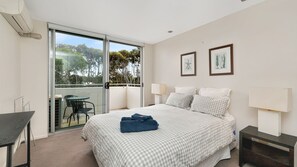 Hotellihuone, jossa on sänky, työpöytä, tuoli, parveke, jolta on näkymä puita kohti, sekä kaksi kehystettyä kuvaa seinällä.