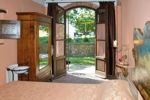 A bedroom with a large bed, a wooden wardrobe, a small table with a chair, and an open door leading to an outdoor area with a table and chairs. - Villa on organic farm between Chianti and Arezzo (Civitella In Val di Chiana)