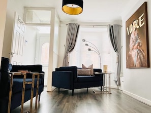A modern living room with a dark blue sofa, a small side table, a floor lamp, and a large window with curtains. - Cozy 2 bed home-15 mins to City/Anfield (Merseyside)
