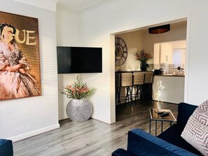 A modern living room with a flat-screen TV, a sofa, and a coffee table. There is a kitchen area visible in the background with bar stools. - Cozy 2 bed home-15 mins to City/Anfield (Merseyside)