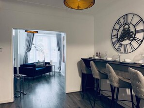 A modern living room with a dark blue sofa, a dining area with a bar, and a large wall clock. - Cozy 2 bed home-15 mins to City/Anfield (Merseyside)