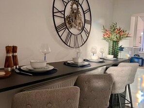 A modern kitchen with a central island, a large wall clock, and a dining area with chairs and a table set for two. - Cozy 2 bed home-15 mins to City/Anfield (Merseyside)