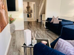 A modern living room with a blue sofa, a small table with a box and a vase, and a staircase leading to a kitchen. - Cozy 2 bed home-15 mins to City/Anfield (Merseyside)