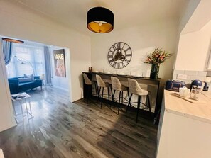 A modern kitchen with a dining area, featuring a clock, bar stools, and a dining table. - Cozy 2 bed home-15 mins to City/Anfield (Merseyside)