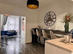 A modern living room with a blue sofa, a dining area with a black table and two chairs, and a large wall clock. - Cozy 2 bed home-15 mins to City/Anfield (Merseyside)