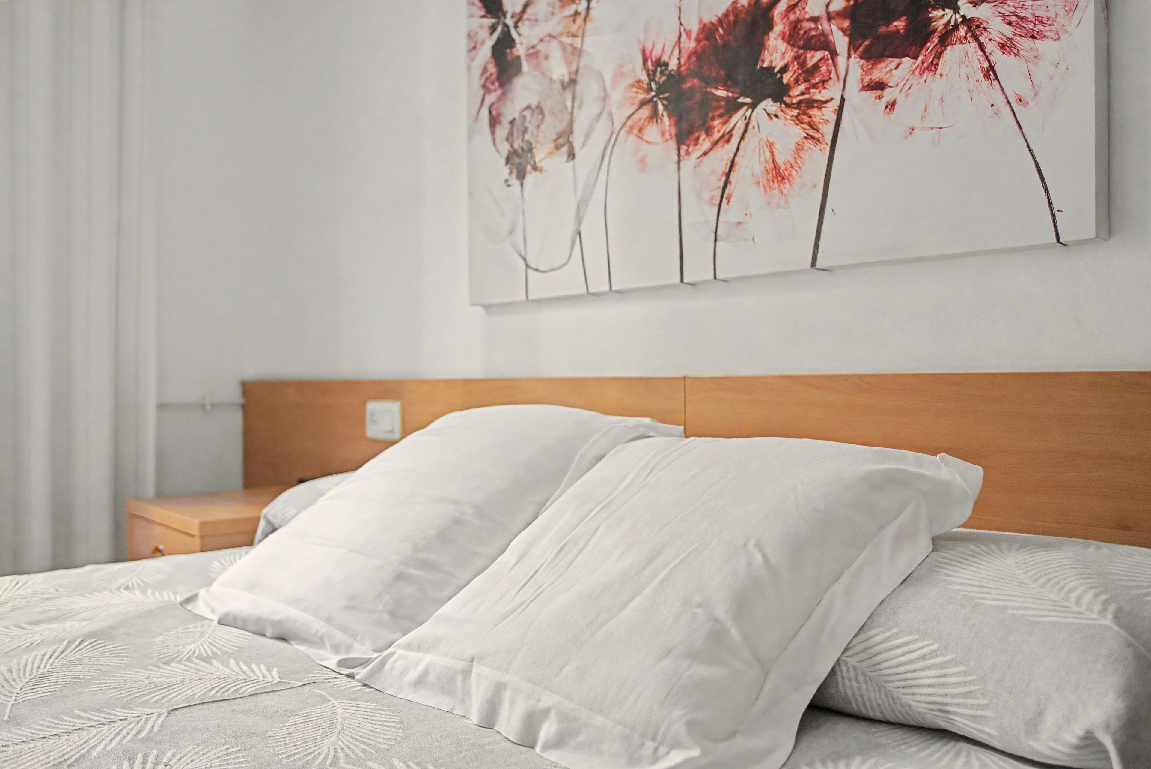 Una cama bien hecha con sábanas blancas y un cabecero de madera, una mesita de noche con una lámpara y una pared con una obra de arte floral.