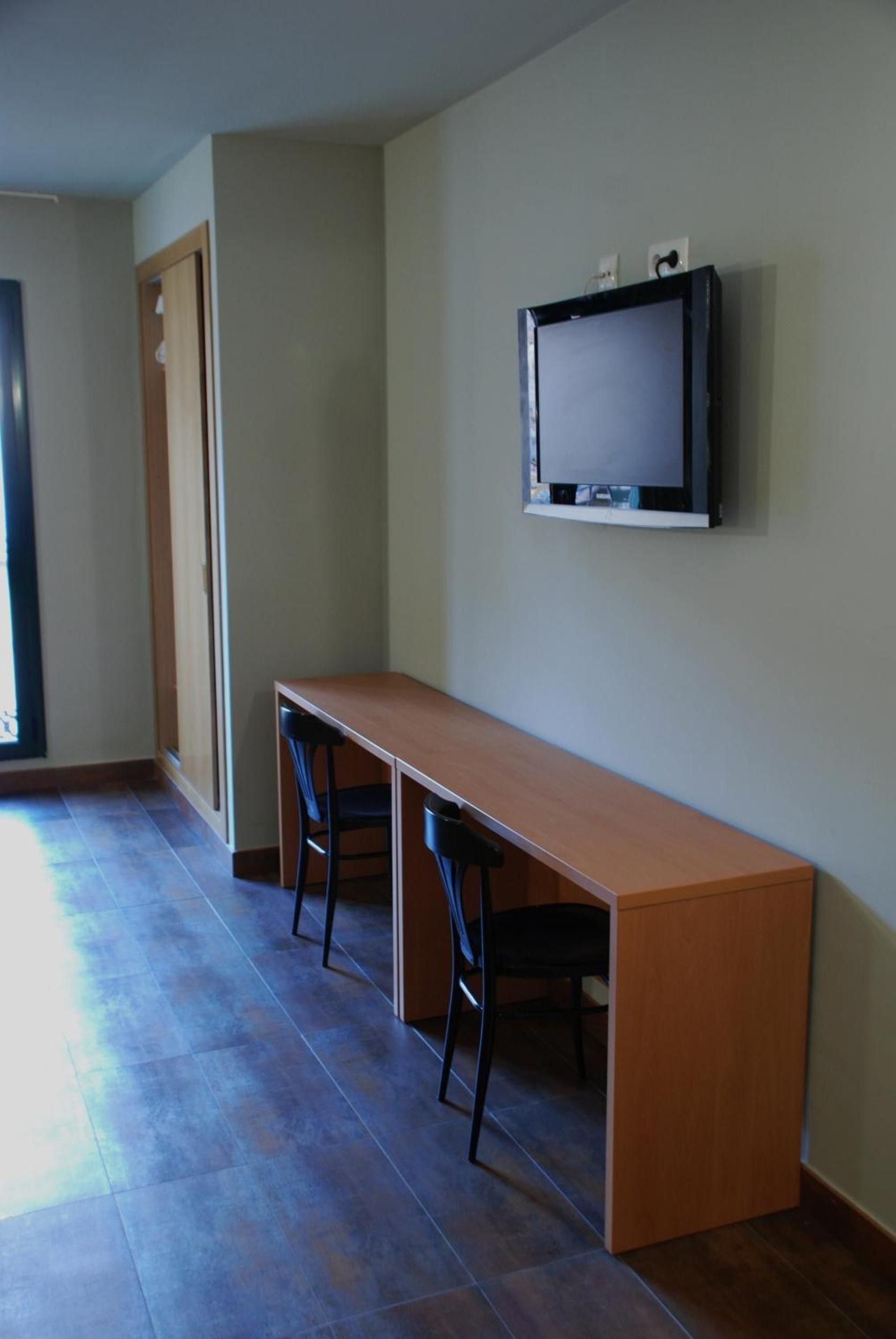Una habitación con una mesa de madera larga, dos sillas, un televisor montado en la pared y una puerta.