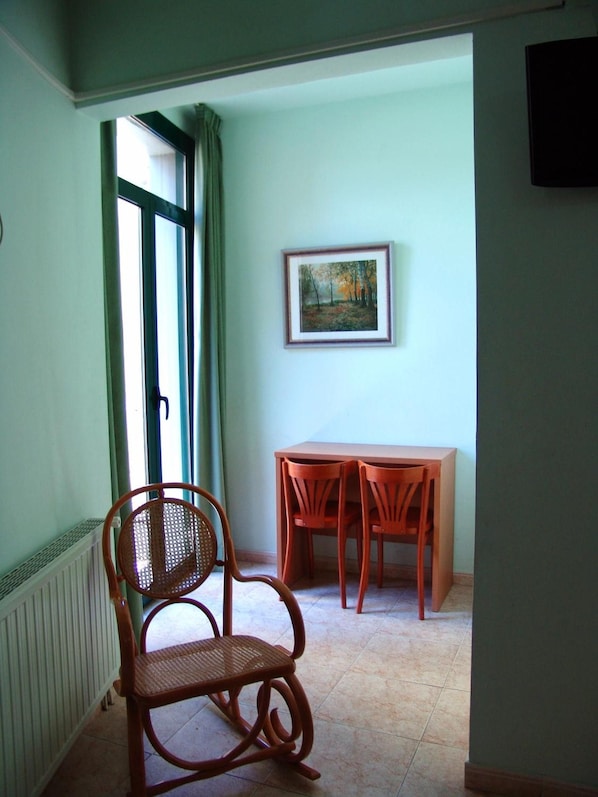 A room with a wicker chair, a table with chairs, a painting on the wall, and a window with curtains.