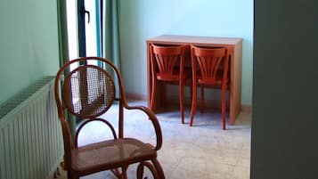 Une pièce avec un fauteuil en osier, une table entourée de chaises, un tableau accroché au mur et une fenêtre avec des rideaux.
