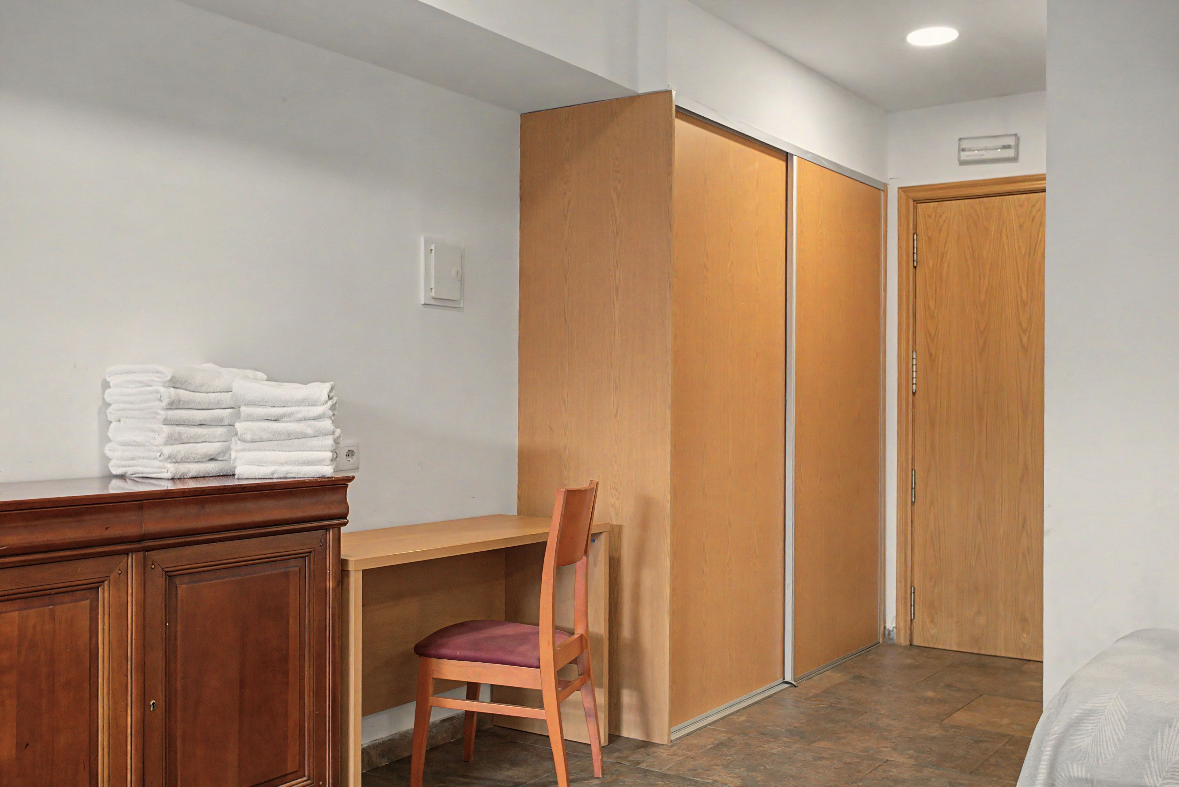 A room with a wooden desk, a chair, a stack of white towels, and a wardrobe.