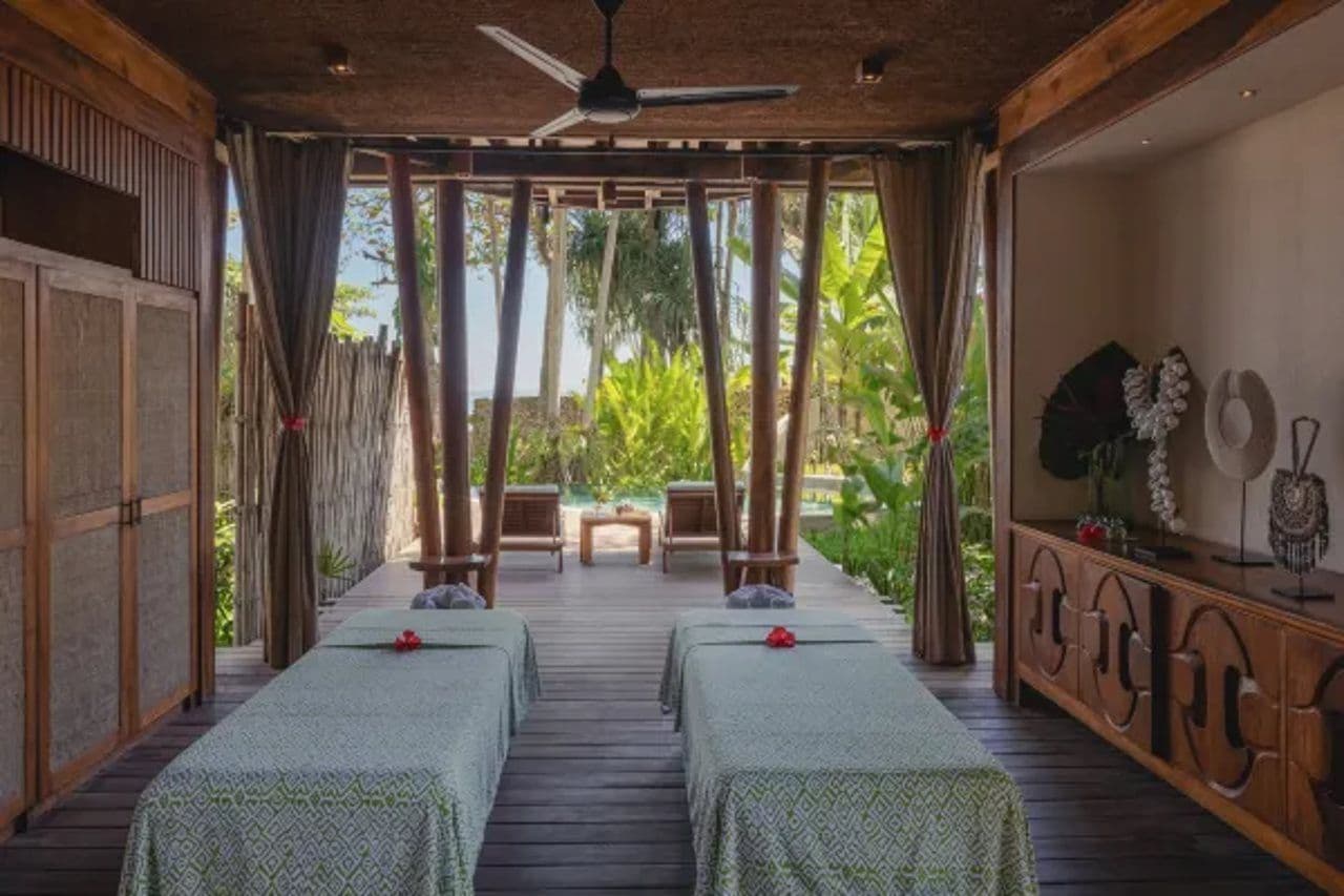 A room with two beds, a ceiling fan, a wooden bench, and a view of greenery outside.