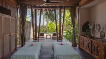 A room with two beds, a ceiling fan, a wooden bench, and a view of greenery outside.