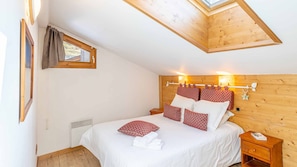 Ein Schlafzimmer mit Holzbalkendecke, einem Oberlicht, einem Bett mit weißer Bettwäsche und roten Kissen, einem Nachttisch und einer an der Wand befestigten Leuchte.