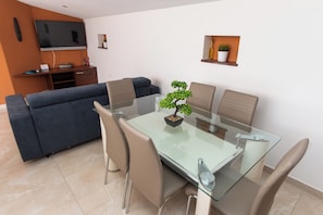 A modern dining area with a glass table, four chairs, a sofa, a TV, and a small shelf with plants. - Penthouse apartment for 4+2 people. Private hot tub on your own terrace. (Podglavica)
