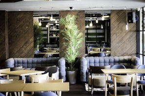 Een modern caféinterieur met lederen zitplaatsen, tafels, hangende lampen en een grote plant in het midden.