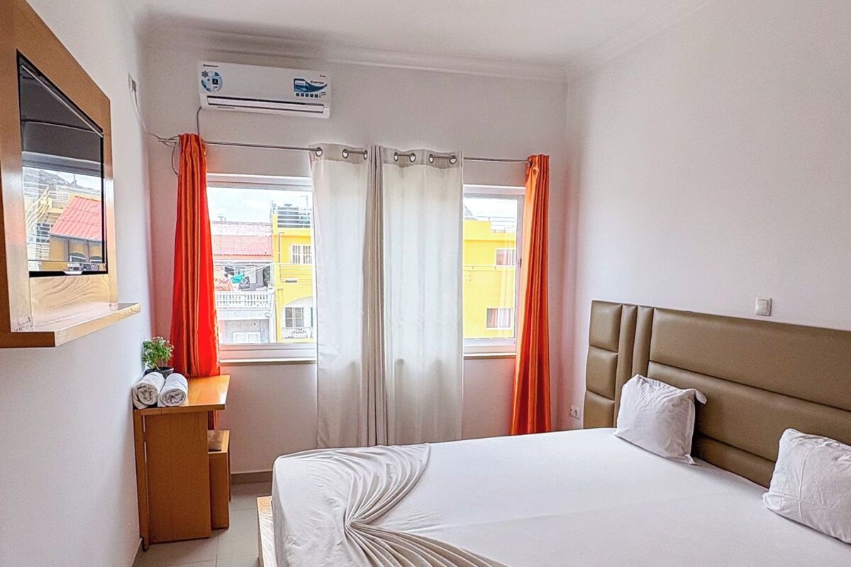 Een hotelkamer met een groot bed, een nachtkastje, een raam met gordijnen en uitzicht op gebouwen.