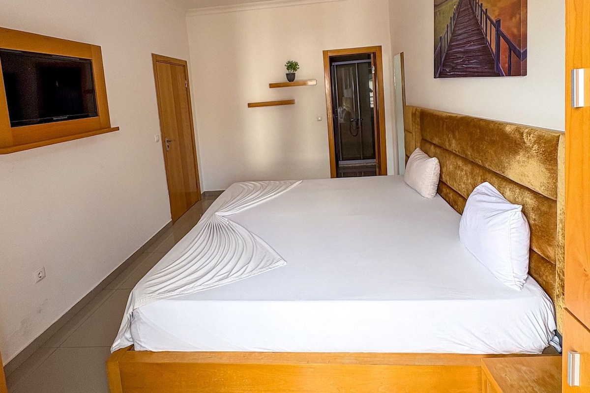 Een slaapkamer met een bed, een schilderij, een deur en een klein plankje met een plant.