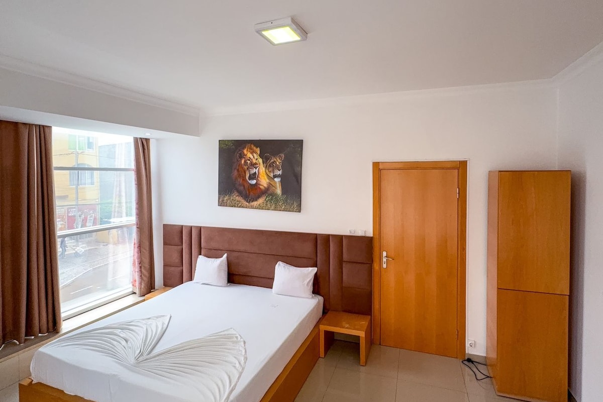 Een hotelkamer met een groot bed, een klein nachtkastje, een kledingkast en een schilderij van leeuwen aan de muur.