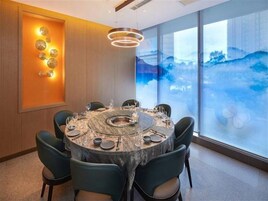 Una mesa de comedor redonda con sillas, una araña y vistas al exterior a través de amplios ventanales.