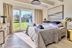Ein Schlafzimmer mit Bett, Nachttisch, Lampe, einem Gemälde und Blick ins Freie.
