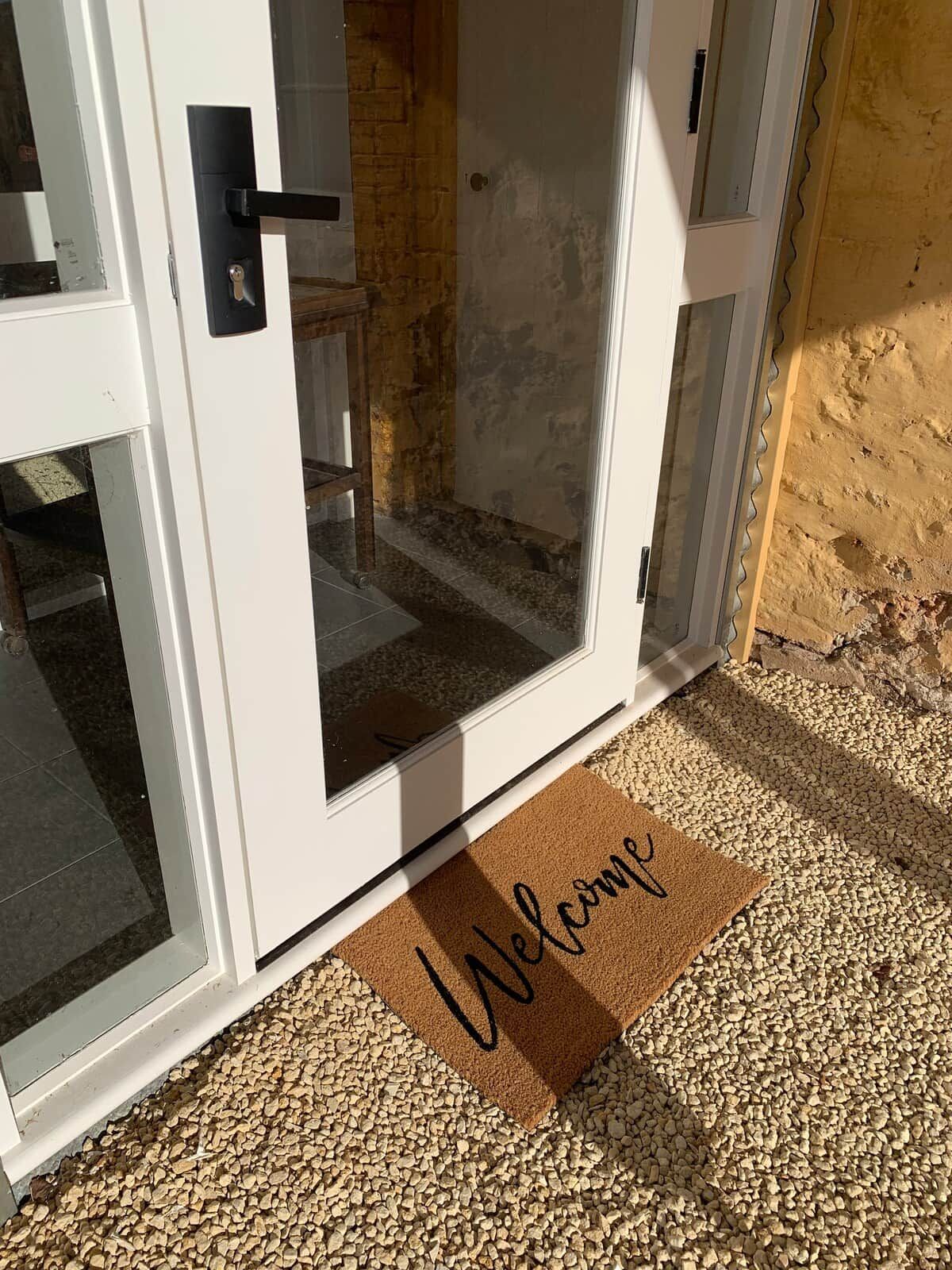 Une porte blanche avec un panneau de verre, un paillasson sur lequel est écrit « Bienvenue », et un chemin de gravier menant à la porte.