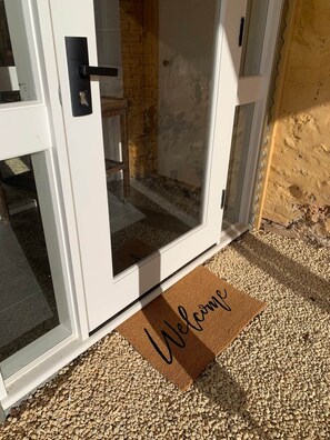Une porte blanche avec un panneau de verre, un paillasson sur lequel est écrit « Bienvenue », et un chemin de gravier menant à la porte.