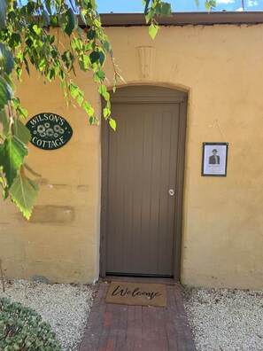 Une porte avec un paillasson sur lequel est écrit « Bienvenue », une pancarte indiquant « Cottage Wilson », et un tableau encadré accroché au mur.
