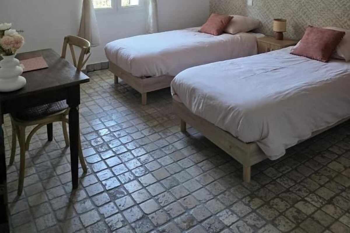 A room with two beds, a table with chairs, a window with curtains, and a wall clock.