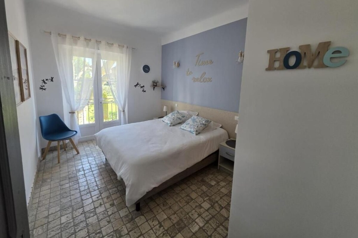 A bedroom with a bed, a chair, a window with curtains, a wall with a 'Home' sign, and a tiled floor.