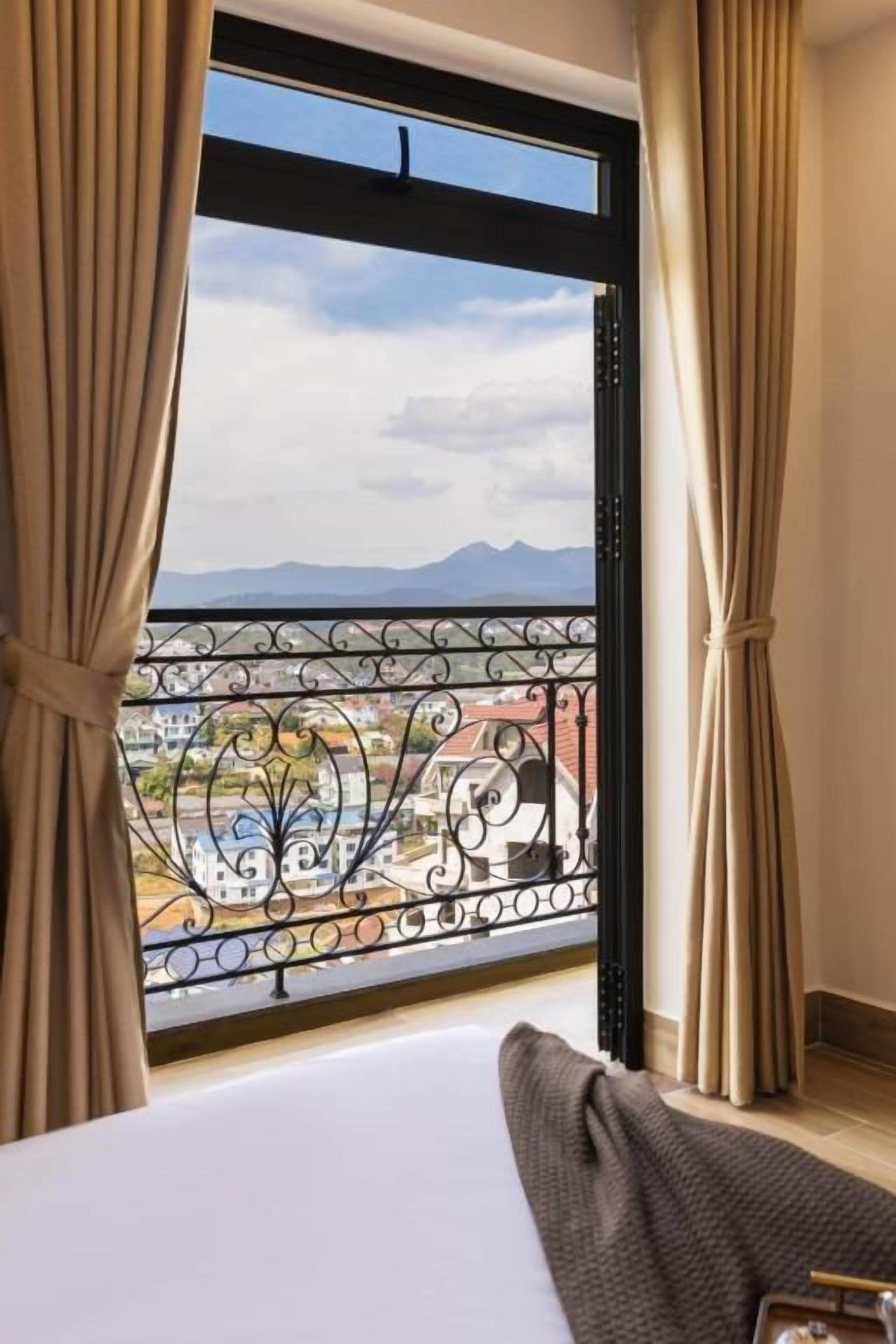 Habitación de hotel con cama, cortinas y vistas a montañas a través de una ventana con baranda de balcón.