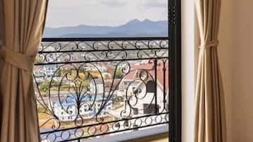 Habitación de hotel con cama, cortinas y vistas a montañas a través de una ventana con baranda de balcón.