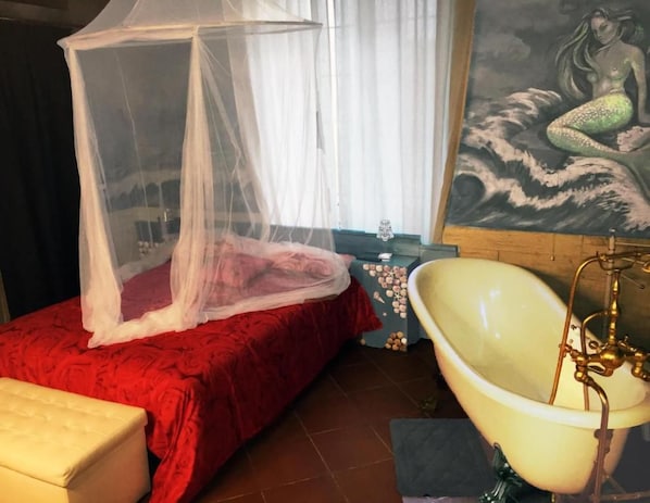 Um quarto com cama sob mosquiteiro, banheira com detalhes em bronze e pintura de sereia na parede.