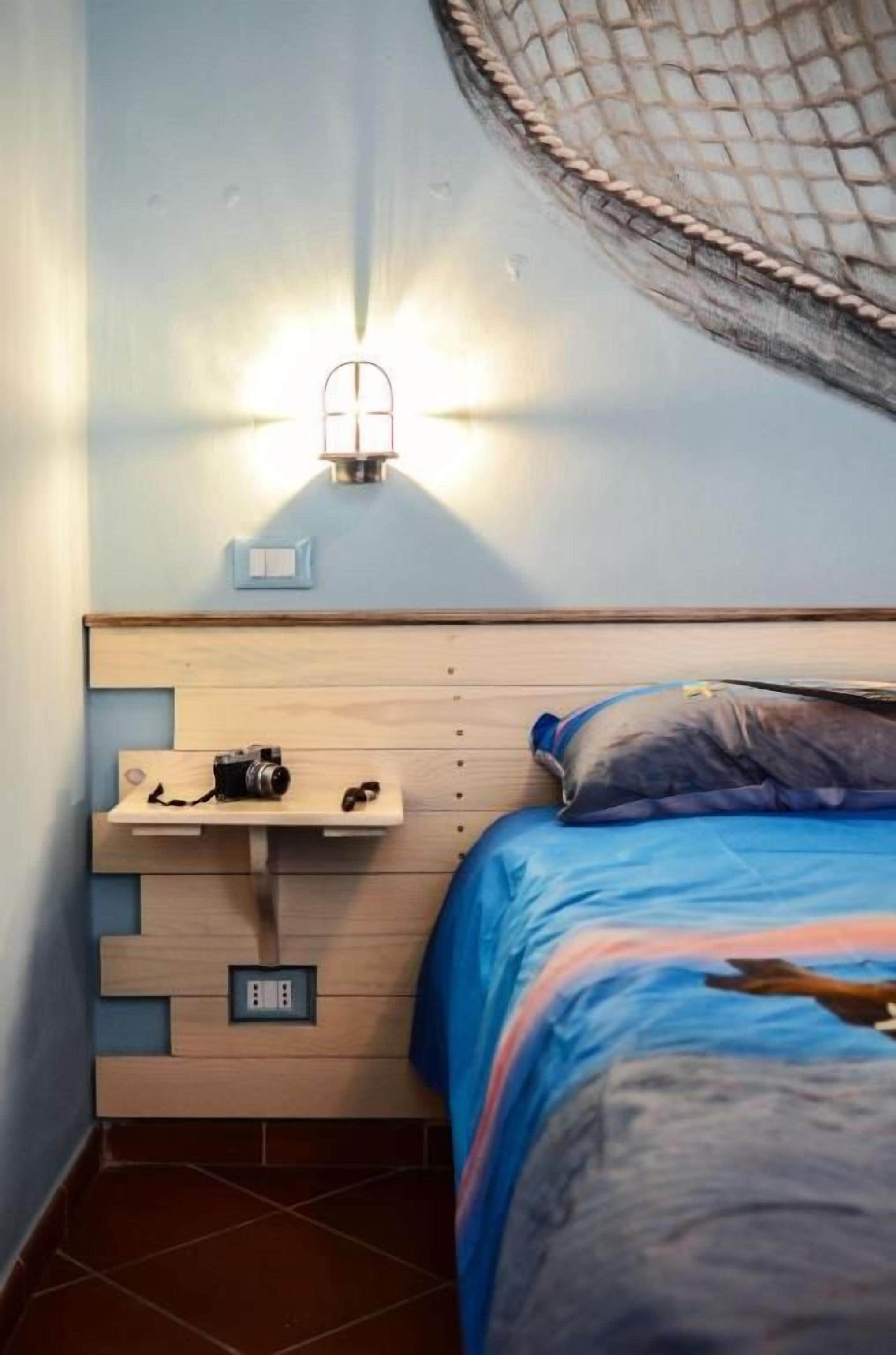 A bedroom with a bed, a nightstand, a camera, and a wall-mounted light fixture.