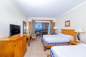 Une chambre d’hôtel avec un lit, un bureau avec une télévision, une chaise et une vue sur la mer par une porte vitrée.