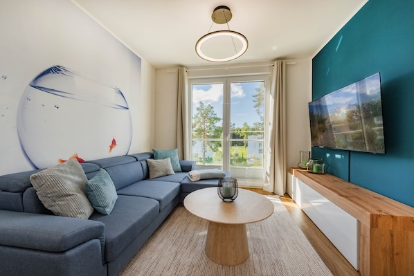 Ein modernes Wohnzimmer mit einer Eckcouch, einem runden Couchtisch, einem an der Wand befestigten Fernseher und einem großen Fenster mit Blick auf Grünflächen.