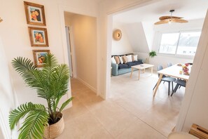 Ein modernes Wohnzimmer mit einer Couch, einem Couchtisch und einem Esstisch. Eine große Topfpflanze und gerahmte Bilder sind an der Wand zu sehen.