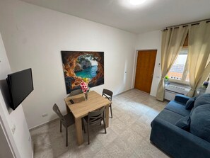 A living room with a blue sofa, a wooden dining table with chairs, a painting of a coastal scene, and a television mounted on the wall. - Holiday Home 'De Rosa'S Home 1' (Tortora Marina)