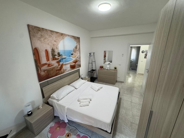 A modern bedroom with a large bed, a nightstand, a dresser, and a painting of a coastal scene.