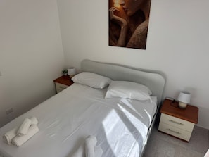 A neatly made bed with white linens and pillows, a bedside table with a lamp, and a small plant.