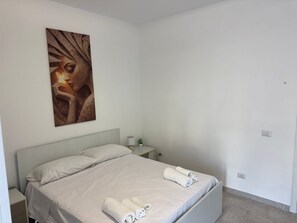 Un dormitorio con cama, mesita de noche con lámpara y un cuadro colgado en la pared.