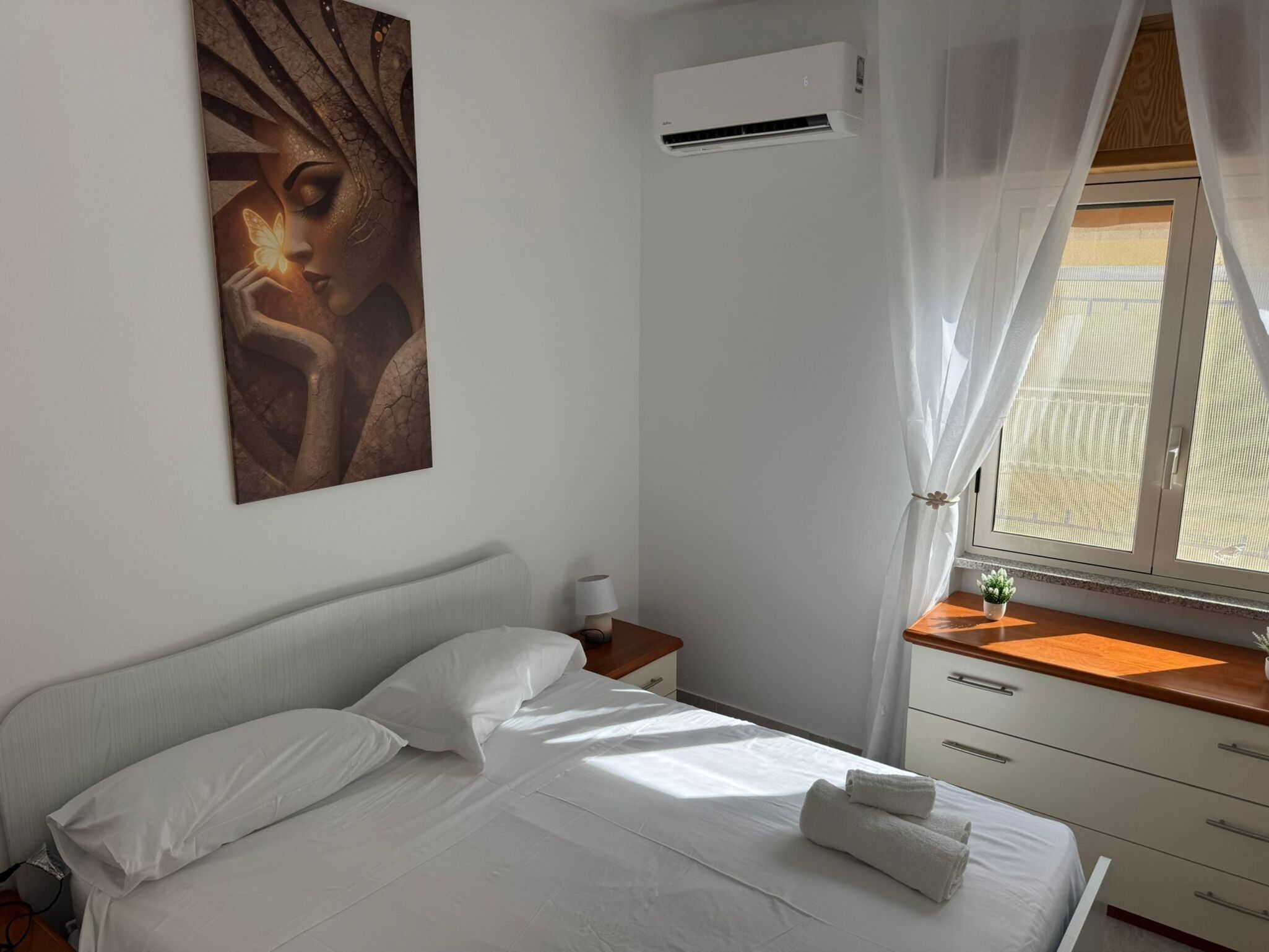 Ein Hotelzimmer mit einem Bett, einem Nachttisch, einer Klimaanlage, einem Fenster mit Vorhängen und einem Wandbild.