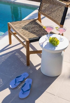 Escena junto a la piscina con una silla de mimbre, una mesita redonda con dos bebidas y una guarnición de frutas, y un par de chanclas en el suelo.