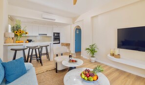 Una sala de estar moderna con un sofá, una mesa de centro con fruta, un televisor montado en la pared y una zona de cocina con una puerta azul.