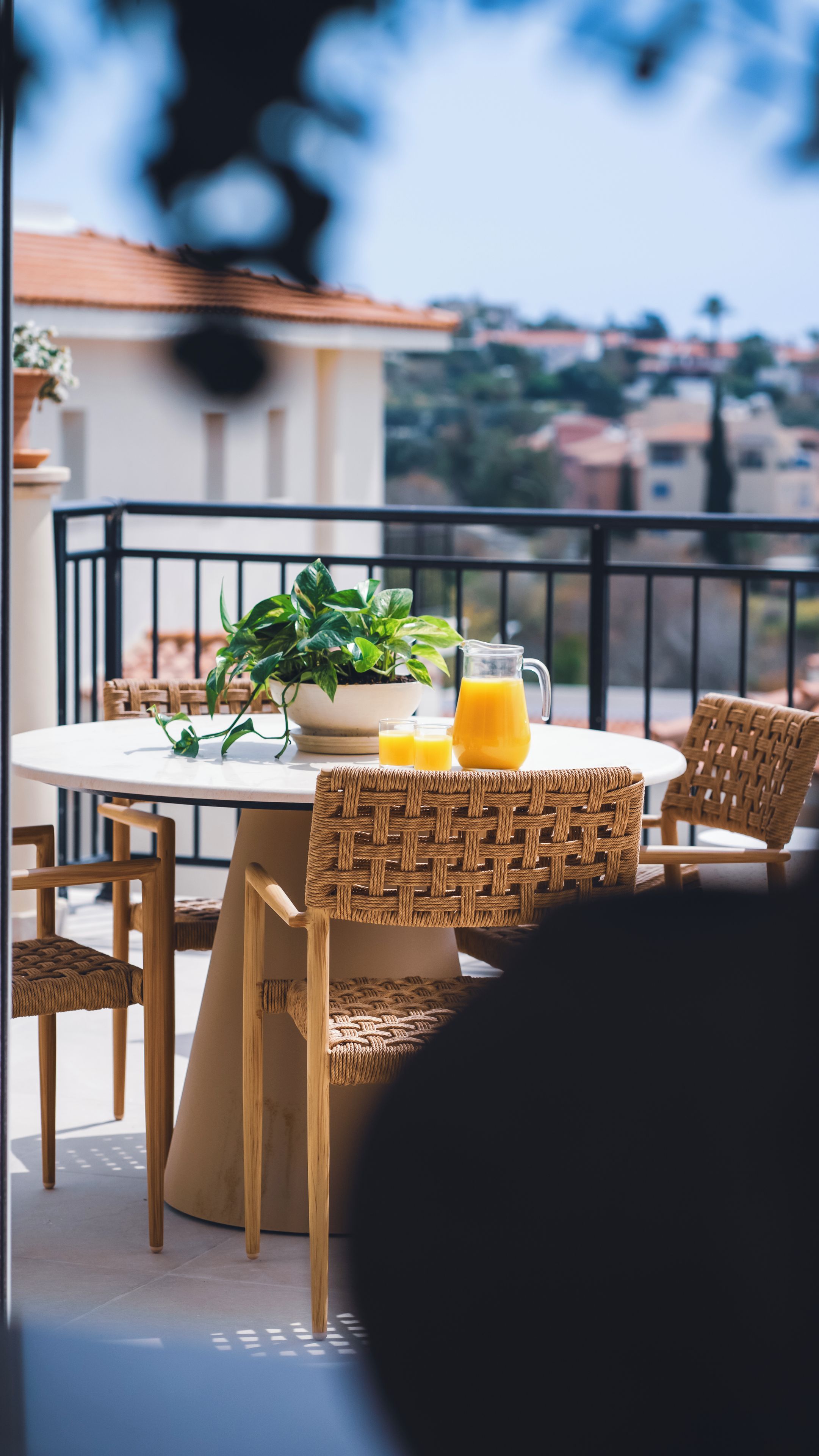 Une table ronde ornée d’un pot de fleurs, d’une carafe de jus d’orange et de deux verres, le tout placé sur un balcon agrémenté de chaises en osier.