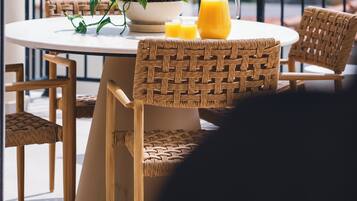 Une table ronde ornée d’un pot de fleurs, d’une carafe de jus d’orange et de deux verres, le tout placé sur un balcon agrémenté de chaises en osier.