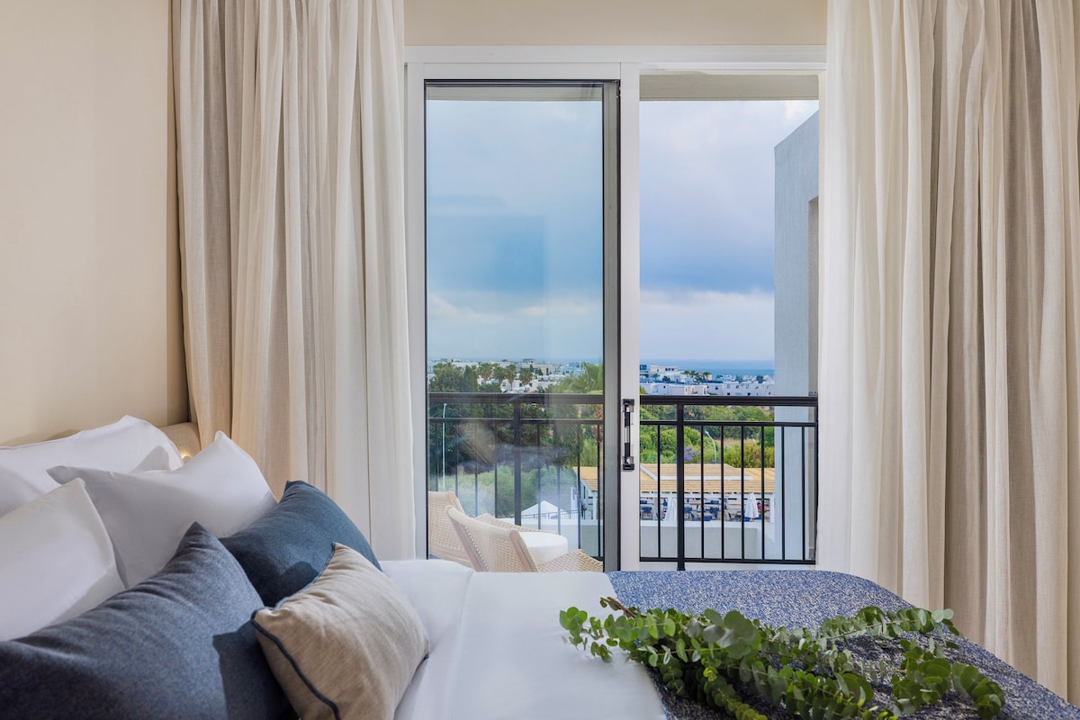 A hotel room with a bed, a balcony with a view of the city, and a dining area with outdoor seating.