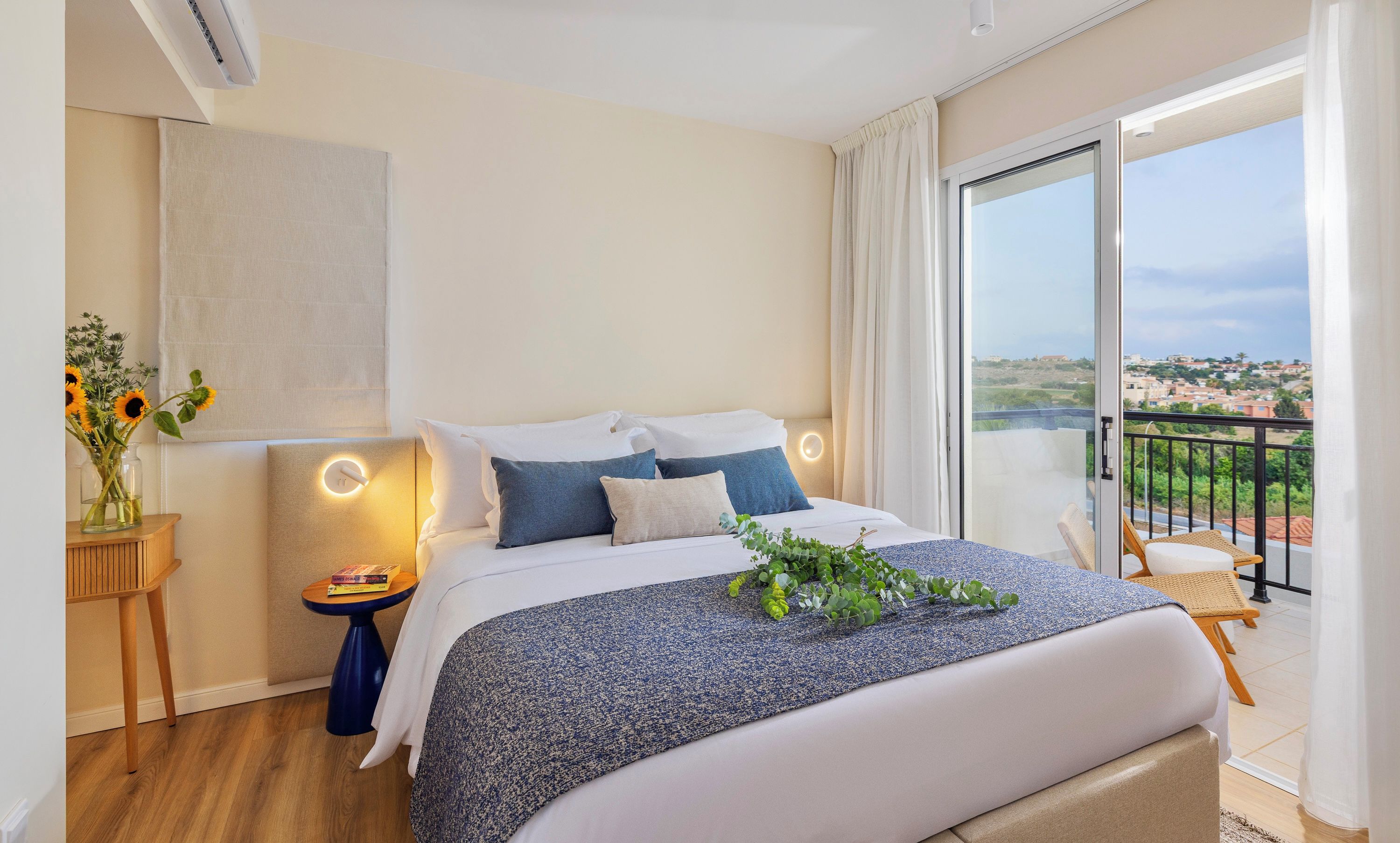 A modern hotel room with a large bed, a small side table, a nightstand, a view of the city through a balcony, and a large window with curtains.