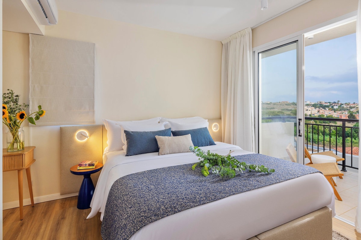 A modern hotel room with a large bed, a small side table, a nightstand, a view of the city through a balcony, and a large window with curtains.