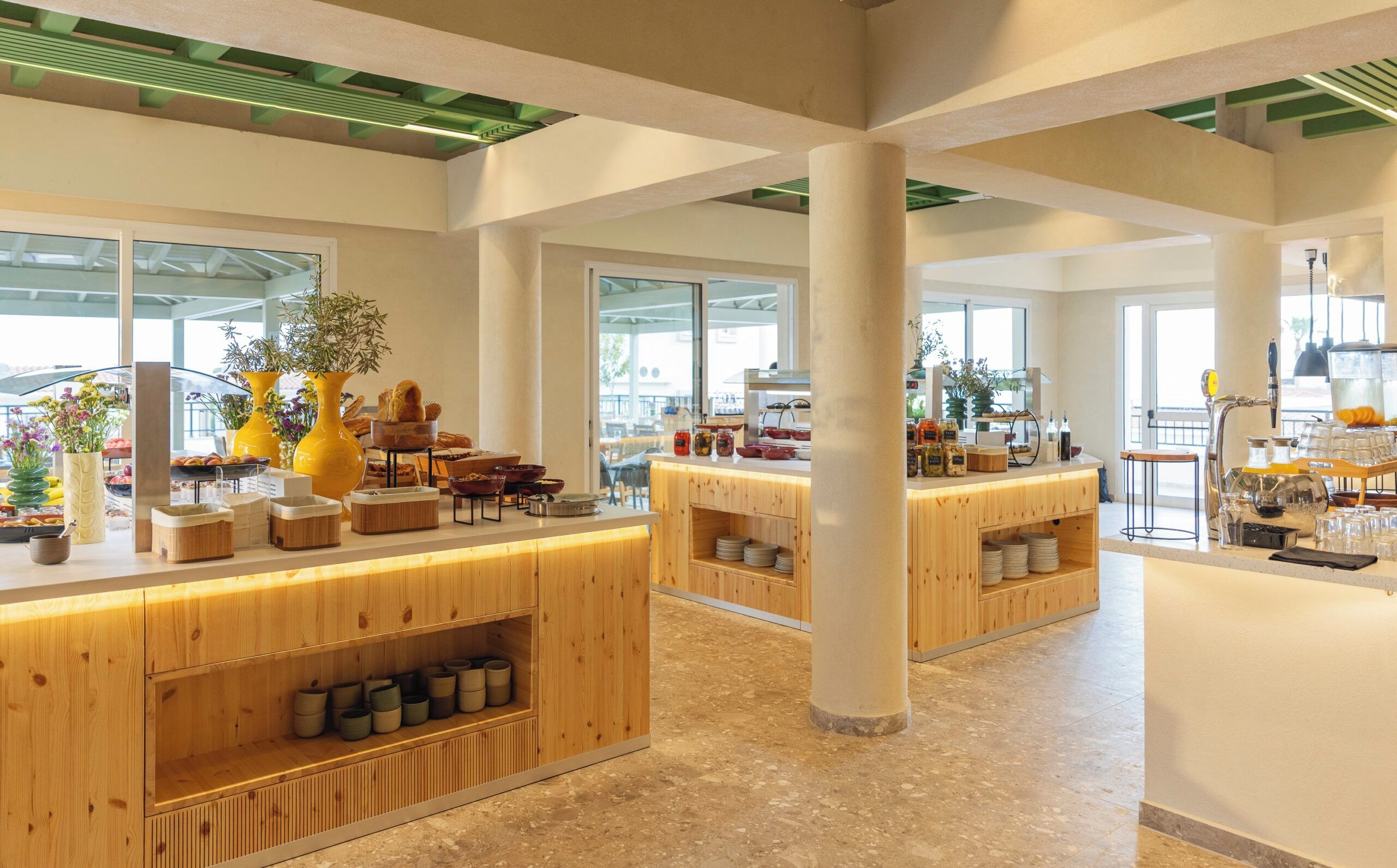 Un hall d’hôtel spacieux, avec des comptoirs d’accueil en bois, une variété de plantes et une vue sur l’extérieur grâce à de grandes fenêtres.